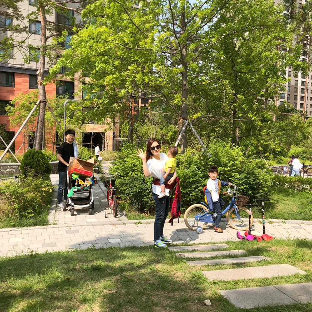 화창한 오후 자이 아파트 단지에서 봄 산책을 즐기는 가족의 모습. 아기를 앉고 활짝 웃고 있는 엄마와 유모차를 잡고 서 있는 아빠, 자전거를 타고 있는 어린 남자 아이 - 자이브랜드매거진-비욘드아파트먼트-동천자이-봄산책