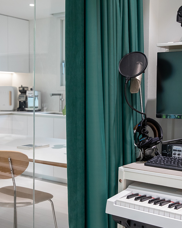 자이매거진-비욘드아파트먼트-은계파크자이-주방 옆 곡면 유리알파룸-곡면 유리로 된 알파룸에 건반과 스피커, 카메라, 모니터 등을 놓고 초록색 커튼을 달아 음악 작업을 하는 뮤직룸으로 꾸몄다. 유리 밖에는 화이트 주방과 테이블 등의 주방이 보인다.
