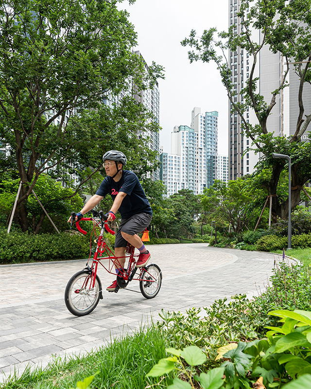 자이매거진-비욘드아파트먼트-서초그랑자이-초록이 우거진 서초그랑자이 아파트 단지에서 헬맷을 쓰고 빨간색 자전거를 타고 있는 중년 남성