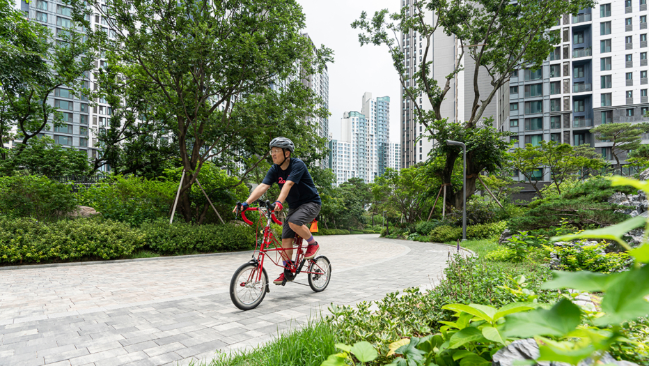 자이매거진-비욘드아파트먼트-서초그랑자이-초록이 우거진 서초그랑자이 아파트 단지에서 헬맷을 쓰고 빨간색 자전거를 타고 있는 중년 남성