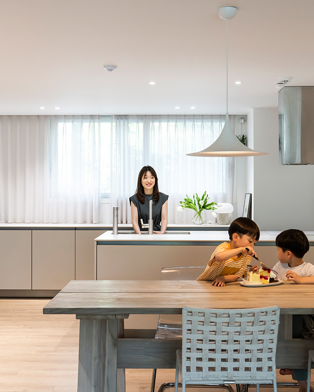 자이매거진-비욘드아파트먼트-수지자이-뉴트럴 컬러로 꾸민 대면형 주방에서 간식을 먹고 있는 아이들과 아일랜드 조리대를 사용 중인 엄마. 상부장을 없애고 커튼을 드리워 개방감이 느껴지는 주방
