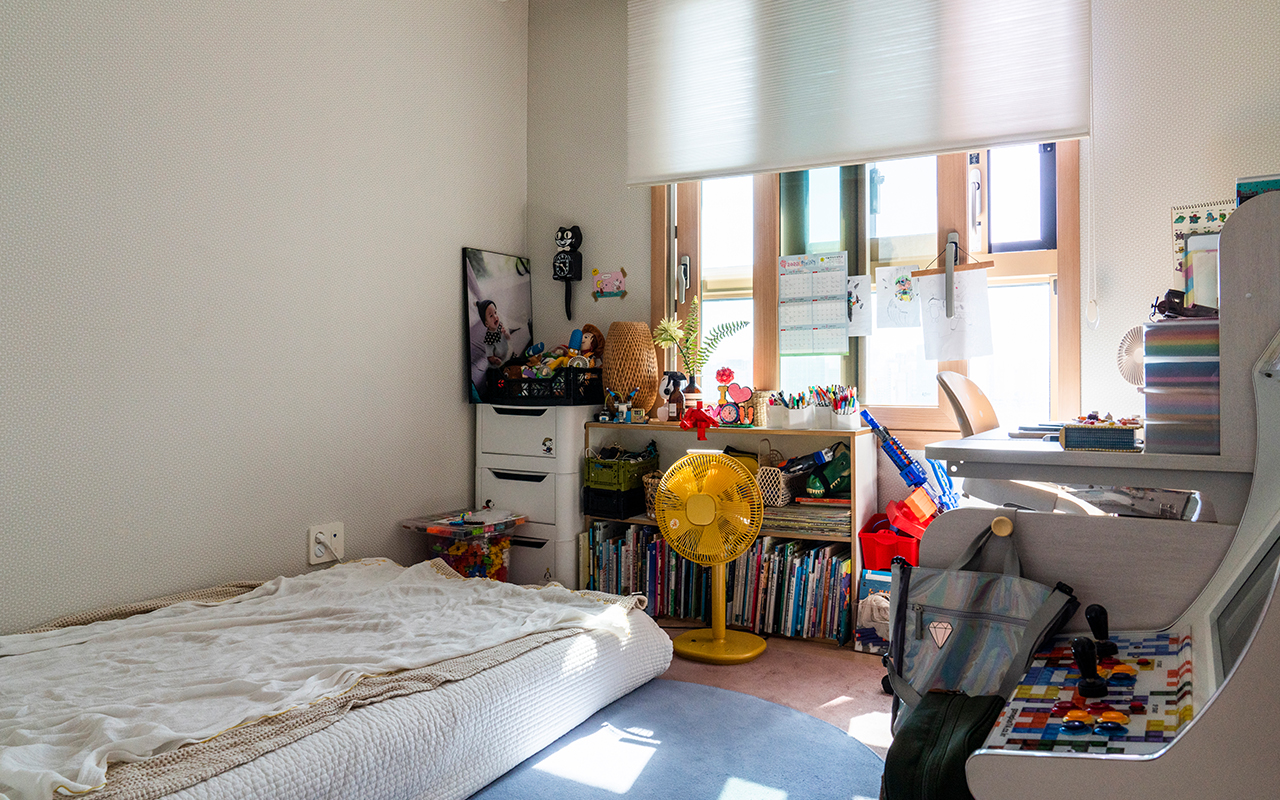 자이매거진-비욘드아파트먼트-침대, 책상, 오락기, 책, 선풍기 등이 있는 작은 방